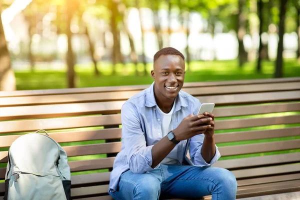 Портрет афроамериканца, использующего мобильный телефон на скамейке в летнем парке — стоковое фото