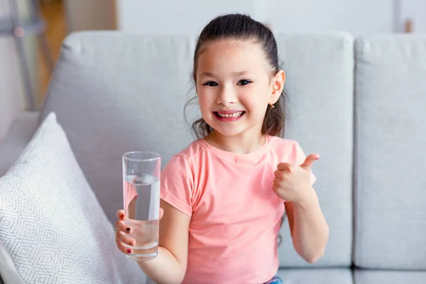 Menina chinesa segurando vidro de água Gesturing Thumbs-Up sentado dentro de casa — Fotografia de Stock