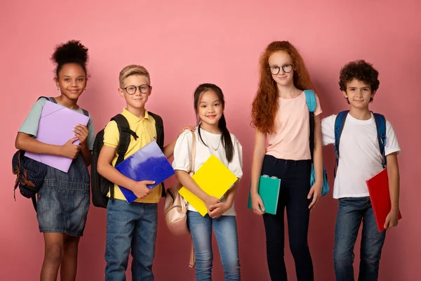Különböző nemzetiségű iskolások jegyzetfüzetekkel és hátizsákkal rózsaszín háttérrel — Stock Fotó