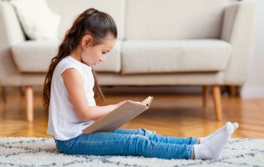 Evdeki halıda kitap okuyan sevimli kız.