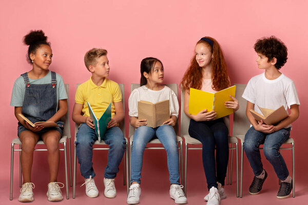 Classmates of different races with school supplies looking at each other on chairs over pink background
