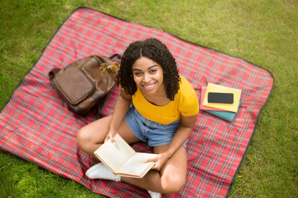 Крута чорна дівчина читає цікаву книгу на пікнік ковдрі в парку, вид зверху — стокове фото