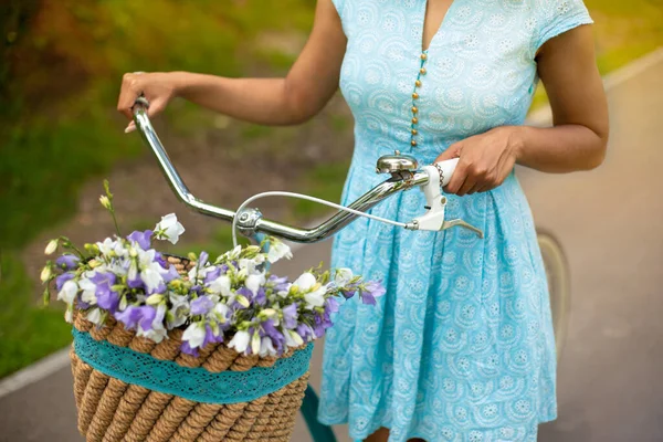 Vista recortada de niña negra sosteniendo bicicleta con cesta llena de flores en el parque, de cerca — Foto de Stock