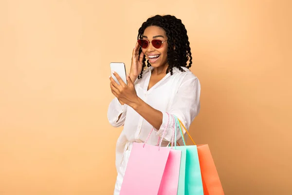 Eccitata nero signora utilizzando telefono holding shopper borse, beige sfondo — Foto Stock