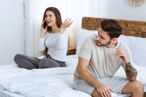 Errores en la relación. Chica alegre hablando en el teléfono inteligente en la cama, marido la mira con disgusto — Foto de Stock