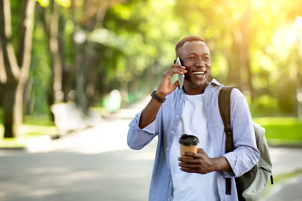 Bonito afro-americano com café para ir falar no smartphone no parque, espaço em branco — Fotografia de Stock