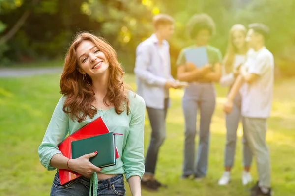 Симпатичная студентка с учебниками, позирующая на улице со своими одноклассниками на заднем плане — стоковое фото