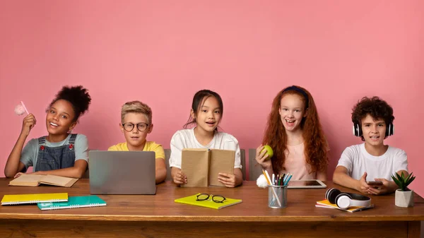 Prêt pour l'école. Divers garçons et filles avec du matériel d'étude et des gadgets assis au bureau, fond rose. Espace libre — Photo