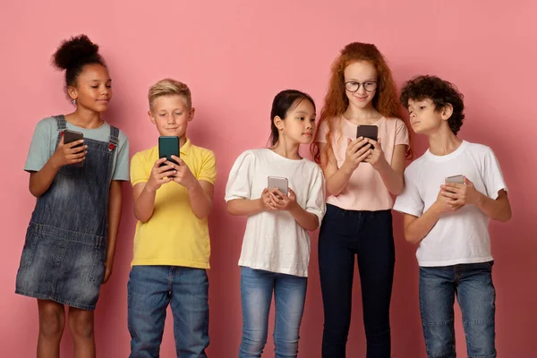 Tecnologías modernas en la escolarización. Niños multiétnicos usando teléfonos celulares juntos sobre fondo rosa — Foto de Stock