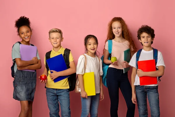 Mi sto preparando per il nuovo anno scolastico. Diversi bambini con zaini, quaderni e frutti di mela su sfondo rosa — Foto Stock