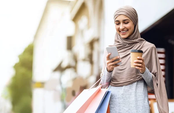 Cheerful arab girl in hijab with shopping bags using phone