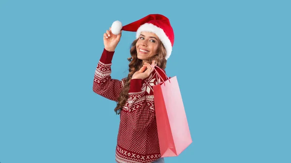 Preciosa mujer joven en jersey de Navidad y Santa sombrero sosteniendo bolsa de compras sobre fondo azul — Foto de Stock
