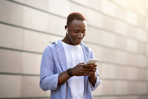 Прохолодний чорний хлопець в навушниках збирає музичний список відтворення на мобільному телефоні біля цегляної стіни на вулиці міста — стокове фото