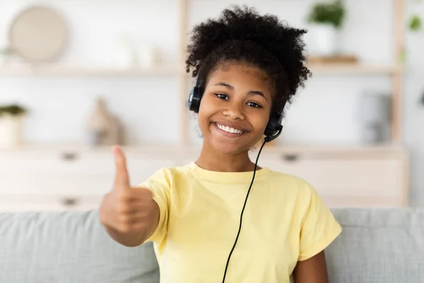 Chica negra con auriculares Gesturing Thumbs-Up sentado en el sofá interior —  Fotos de Stock