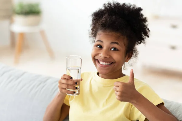 Africana adolescente chica celebración vaso de agua gestos pulgares-up interior —  Fotos de Stock