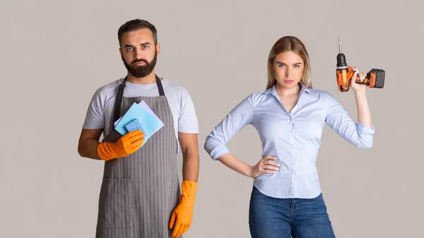 Gender stereotypes and non female profession. Serious man in apron and rubber gloves holding sponges and woman holding a drill — Stock Photo, Image
