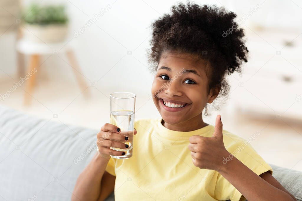 African Teen Girl Holding Glass Of Water Gesturing Thumbs-Up Indoor