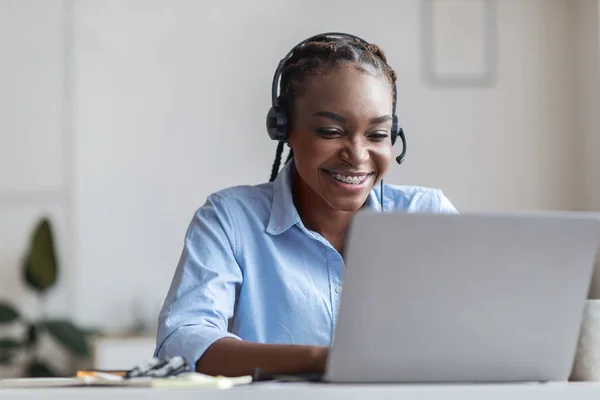 Serviço de suporte de linha direta. Mulher africana sorridente no fone de ouvido trabalhando no escritório moderno — Fotografia de Stock