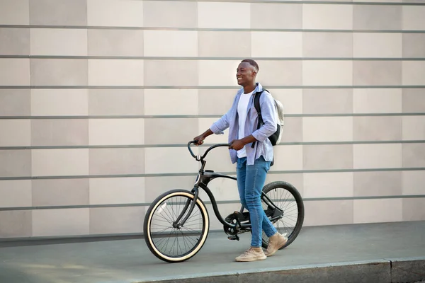 Bonito homem negro com mochila e bicicleta andando perto da parede de tijolo ao ar livre, espaço livre — Fotografia de Stock