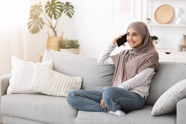 Wanita arab yang bahagia berbicara di telepon genggam sambil duduk di sofa di rumah — Stok Foto