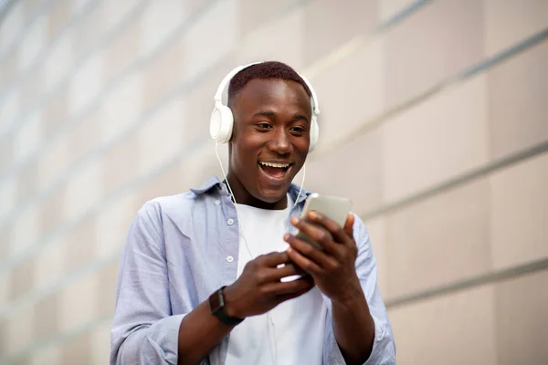 Opgewonden Afro-Amerikaanse man in hoofdtelefoon op zoek naar zijn smartphone scherm in de buurt van bakstenen muur — Stockfoto