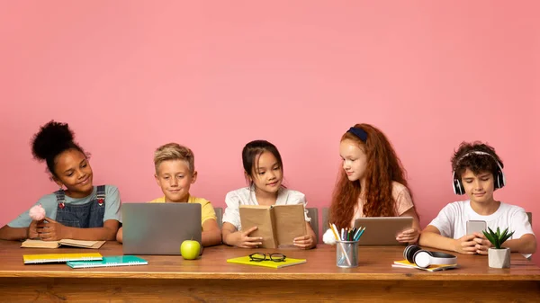 Hora da escola. Crianças multiétnicas com diferentes gadgets e livros sentados à mesa contra fundo rosa, espaço vazio — Fotografia de Stock
