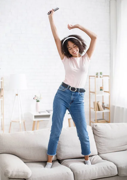 Menari dengan headphone dan bersantai di rumah. Wanita bahagia dengan smartphone di sofa di ruang tamu — Stok Foto