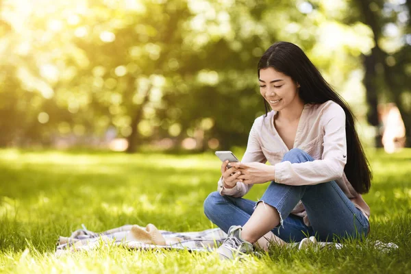 Şehir parkında dinlenirken akıllı telefondan mesaj atan neşeli Asyalı kadın.