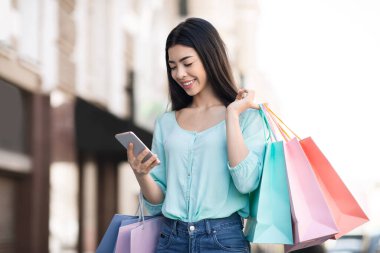 Güzel Asyalı kadın dışarıda alışveriş torbalarıyla dolaşırken akıllı telefonunu kullanıyor.