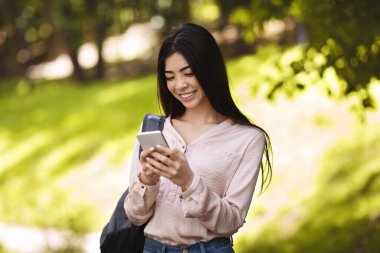 Sırt çantalı neşeli Asyalı genç kız dışarıda akıllı telefon kullanıyor.