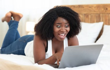 Şaşırmış siyah kız, dizüstü bilgisayar kullanarak yatakta yatıyor.