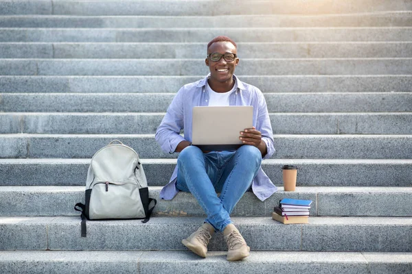 Educação online. Homem negro inteligente com laptop e pilha de livros sentados nas escadas do centro da cidade — Fotografia de Stock