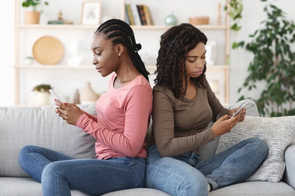 Black girls sitting back to back, using smartphones