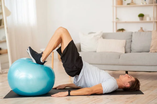 Athletic guy in sportswear and fitness tracker doing abdominal exercises in living room interior — Stock Photo, Image