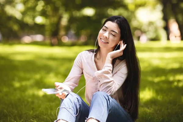 Χαλαρωμένη Ασιάτισσα που απολαμβάνει να ακούει μουσική στο Smartphone Outdoors, φορώντας ακουστικά — Φωτογραφία Αρχείου