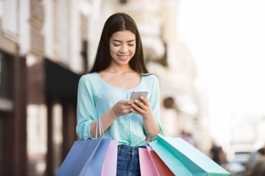Alışveriş merkezinde başarılı bir alışveriş yaptıktan sonra akıllı telefonu açık havada kullanan genç Asyalı kız.