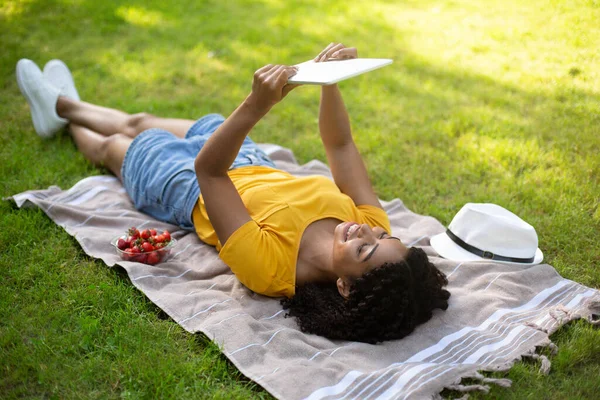 Весела афроамериканська дівчина з планшетом дивиться навчальне відео, лежачи на ковдрі в парку. — стокове фото