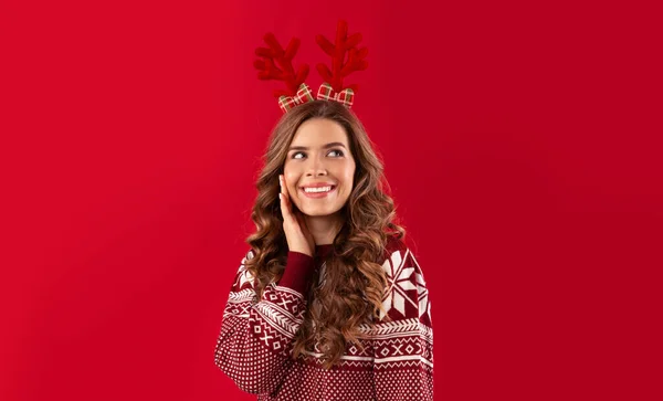 Es Navidad. Hermosa joven en suéter cálido colorido con cuernos de ciervo sobre fondo rojo — Foto de Stock