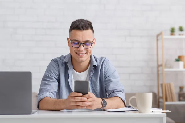 Geben Sie Ihr Telefon ein und arbeiten Sie online. Lächelnder Typ im Hemd blickt aufs Smartphone, sitzt mit Laptop am Tisch — Stockfoto