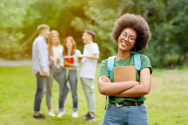 Студенческая жизнь. Улыбающаяся черная девушка позирует перед своими друзьями из колледжа в кампусе — стоковое фото