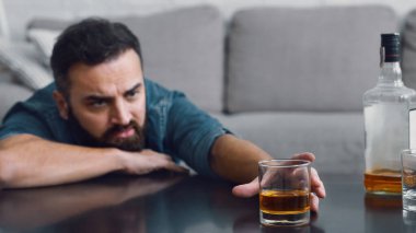 Hüsrana uğramış sakallı adam oturma odasındaki masadan bir bardak viski alır.