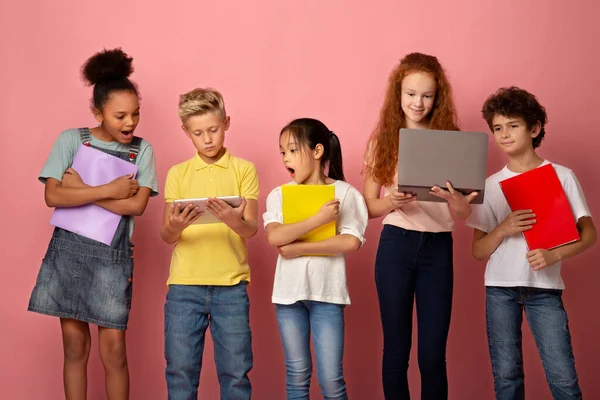 Портрет багатоетнічних школярів з блокнотами, що використовують планшет і ноутбук для навчання на рожевому фоні — стокове фото