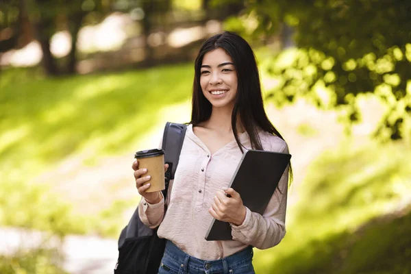 Χαμογελαστή Ασιάτισσα μαθήτρια με σακίδιο και φορητό υπολογιστή που απολαμβάνει τον καφέ απ 'έξω — Φωτογραφία Αρχείου