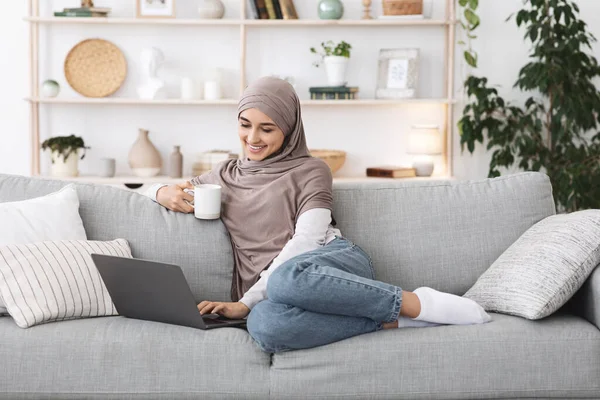 Mujer árabe positivo relajarse con el ordenador portátil y café en el sofá en casa — Foto de Stock