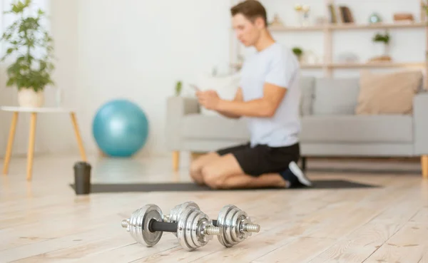 Athlete typing in smartphone, sitting on floor with glass of protein shake, with ball and dumbbells in interior of living room — Stock Photo, Image
