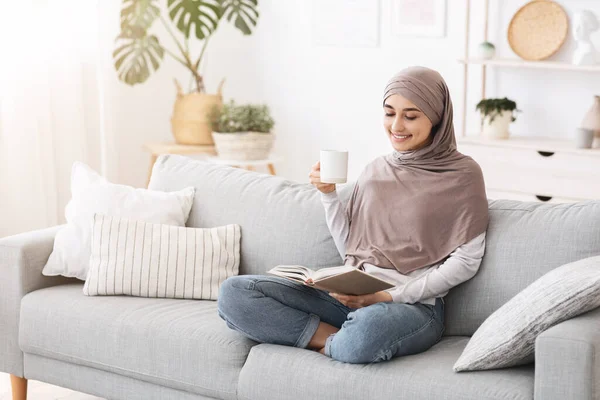 Boldog arab háziasszony lazít a könyv és a kávé a kanapén otthon — Stock Fotó