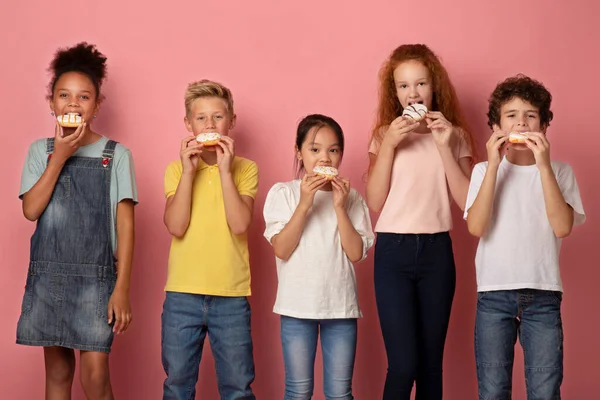 Lindos niños diversos comiendo rosquillas deliciosas sobre fondo rosa — Foto de Stock