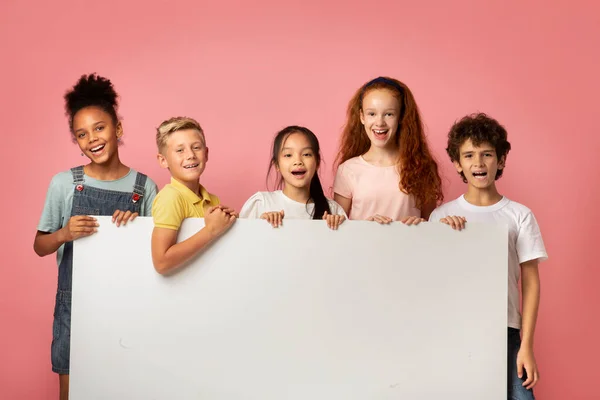 Vrolijke diverse schoolkinderen houden blanco banner met ruimte voor design over roze achtergrond — Stockfoto