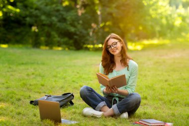 Temiz hava üzerinde çalışıyorum. Neşeli öğrenci kız elinde kitapla çimlerde oturuyor.