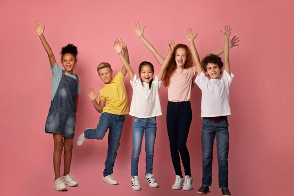 Fröhliche multiethnische Schulkinder heben ihre Hände auf rosa Hintergrund — Stockfoto